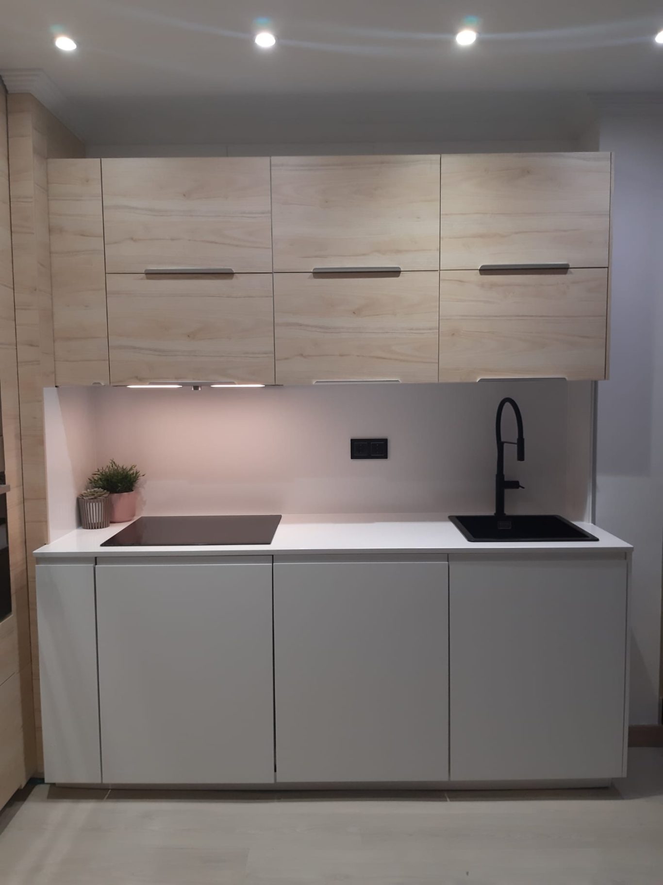 Cocina moderna con armarios de madera clara y encimera blanca, con fregadero negro.